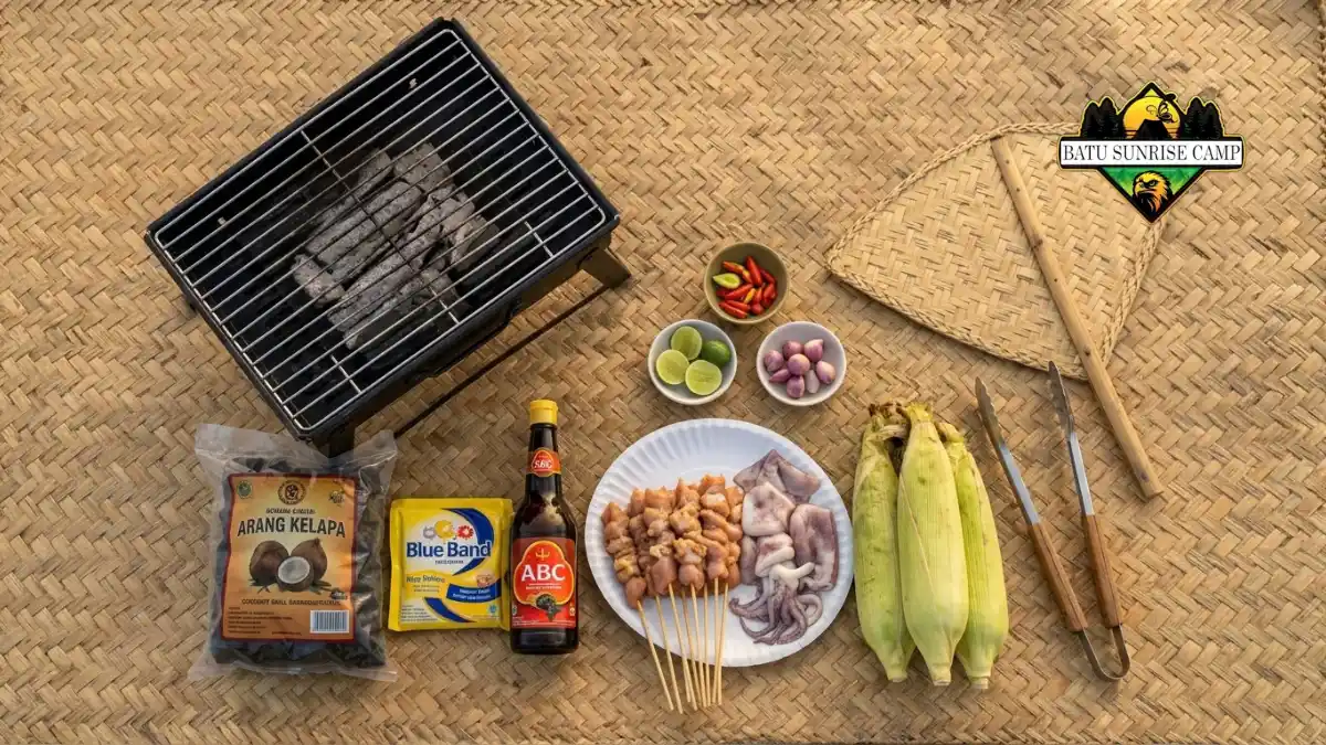 Flat lay checklist alat dan bahan BBQ camping berupa grill portable, arang kelapa, mentega, kecap manis, sate ayam, cumi, jagung, cabai, dan jeruk nipis di atas tikar anyaman