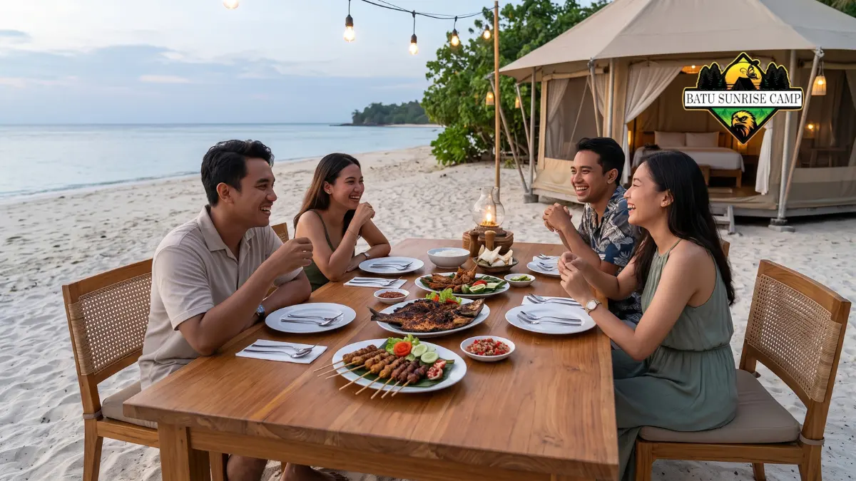 Sekelompok teman sedang menikmati makan malam dan ikan bakar di meja kayu di tepi pantai pasir putih di depan tenda glamping saat senja