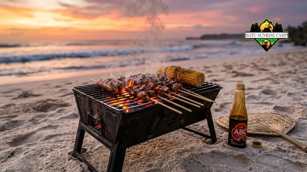 Panggangan portable berisi sate dan jagung bakar di atas pasir pantai saat senja dengan botol kecap dan kipas bambu di sampingnya