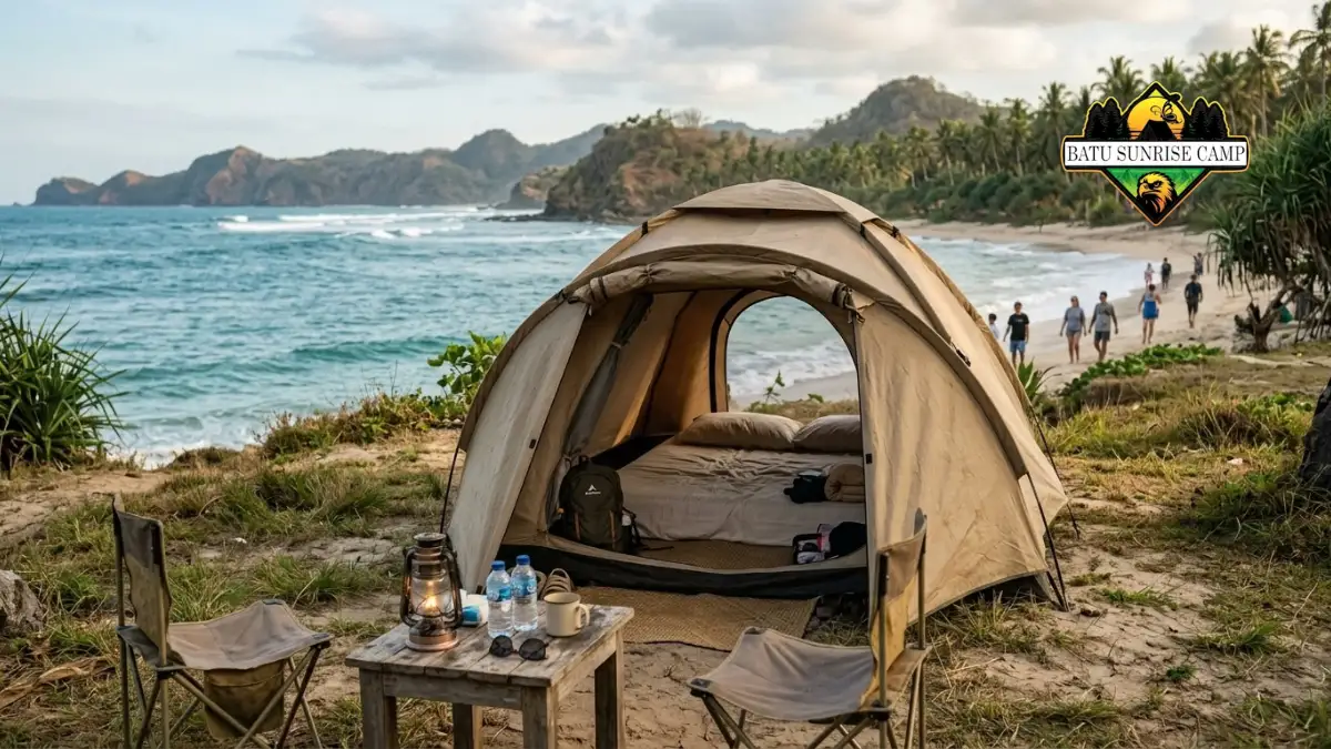 Tenda glamping sederhana dengan kasur dan kursi lipat di area pesisir pantai berpasir dengan pemandangan laut dan perbukitan hijau